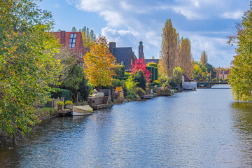 Fototapeta premium A scenic view of a lush garden and traditional brick houses by the water on the Northern Amstel Canal, surrounded by autumn foliage and clear blue sky reflections. Amsterdam, the Netherlands.