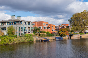 A scenic view of a lush garden and traditional brick houses by the water on the Northern Amstel Canal, surrounded by autumn foliage and clear blue sky reflections. Amsterdam, the Netherlands.