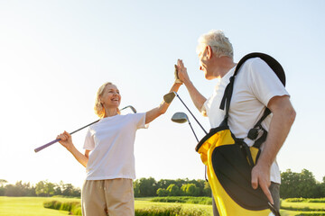 elderly senior couple in uniform celebrating victory and success in golf game and giving high five, old man and woman playing golf on golf course at sunset and doing outdoor sports - Powered by Adobe