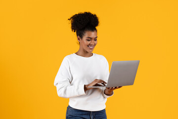 A cheerful young woman stands smiling while using her laptop against a vibrant yellow background. Her relaxed attire suggests she is engaged in casual work or leisure activities. © Prostock-studio