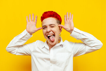 young short-haired red-haired girl fooling around and making faces on a yellow isolated background, hipster woman with piercing shows grimaces and tongue