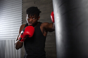 young strong african american boxer in red gloves hits punching bag, muscular athlete trains punch...