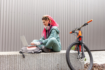young hipster girl with dreadlocks in warm clothes uses a laptop near a bicycle on a background of a gray wall, student cyclist works online in cold weather in autumn