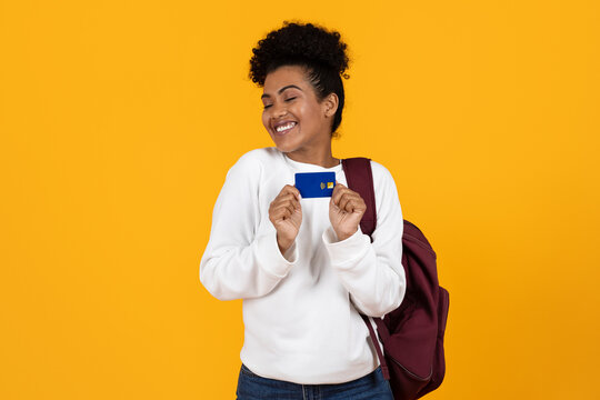 A cheerful young woman in a white sweater and jeans happily displays her credit card while standing against a vibrant yellow backdrop, embodying excitement and financial empowerment.