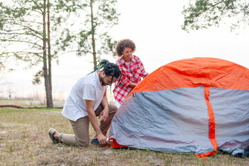 young african american couple hikers pitching tent in forest near lake, man and woman with camping equipment and backpacks traveling and preparing camping site
