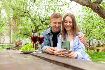 Naklejka premium young couple in love in denim clothes sitting in the garden at a wooden table drinking wine with pork and using a smartphone in the summer, a guy and a girl on a picnic celebrating and taking selfies