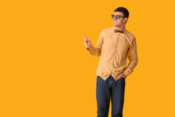 Young man with stylish bow tie and sunglasses pointing at something on yellow background