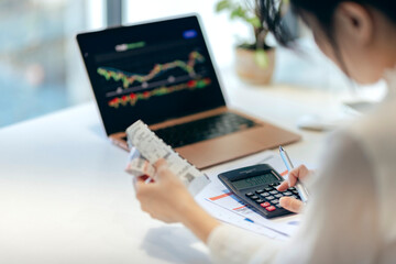 A mother in her home office reviews annual expenses and taxes, with a stock chart displayed on her...