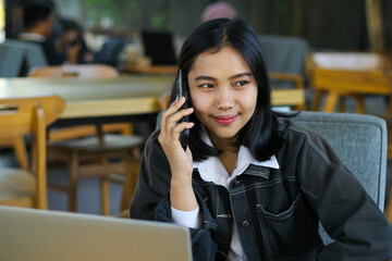 happy asian woman make mobile phone call while working in indoor cafe with laptop, looking away