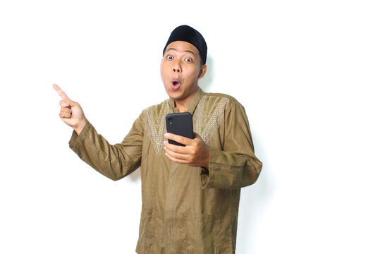 shocked asian muslim man holding mobile phone pointing to beside isolated on white background