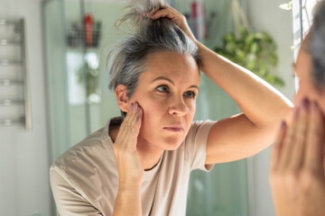 European white-skinned middle-aged woman looks anxiously in the bathroom mirror. Gray hair, wrinkles