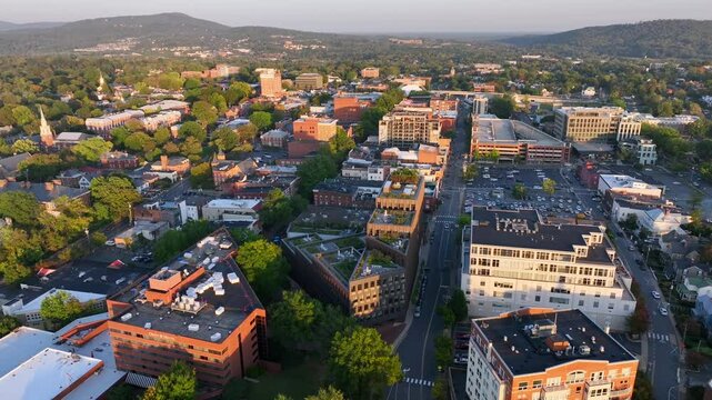 Downtown streets with historic buildings surrounded by trees and hills. Parking lots and rooftops fill urban landscape. Aerial view of Charlottesville, Virginia.