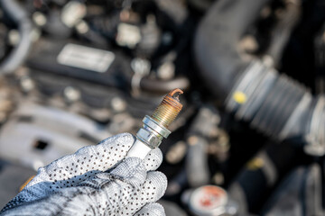 Worn spark plug on hand, and a car engine in the background.