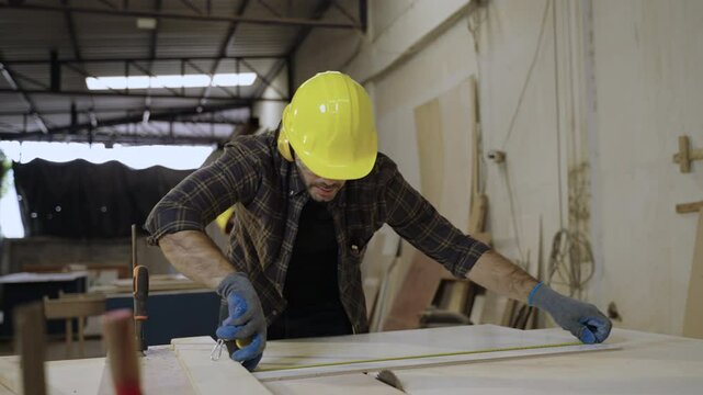 A handsome carpenter man in safety goggles and a helmet standing using the electrical circular saw to cut plank timber in the workplace. Young male carpenter worker working in a furniture factory. 