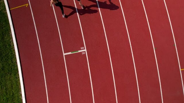 Two athletes preparing on the starting lines of a red running track, captured from an aerial perspective under bright sunlight