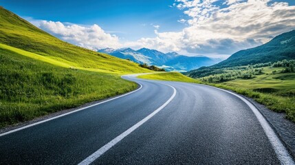 Naklejka premium A winding asphalt road snakes through lush green hills with snow-capped mountains in the background under a clear blue sky with puffy clouds.