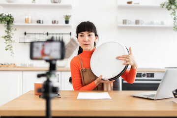 Caucasian female blogger reviews new model of robot vacuum cleaner online. She sits in modern kitchen, engaging with her audience through smartphone mounted on tripod.