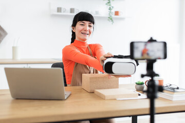 Caucasian female in her twenties unboxing virtual reality headset during online recording session. Enthusiastic expression while showcasing new technology product to audience.