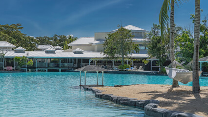 The recreation area by the swimming pool. Gazebo by aquamarine water. The hammock is suspended from the trunks of palm trees. Cottages among the green vegetation in the distance. Blue sky, clouds. 