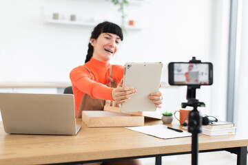 Caucasian female blogger in orange sweater excitedly unboxing new digital tablet during livestream. Displaying product to camera on tripod, sitting at desk with laptop and notepad.