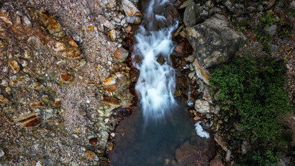 This aerial photo with slow speed features a stream flowing between brown and gray rocks. There are green plants on the right side, creating a contrast with the rocks' colors.