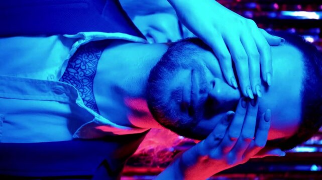 Vertical video. Xmas surprise. Color light couple. Christmas party. Happy woman playing peekaboo with man in blue red pink neon glow on shiny tinsel.