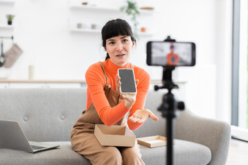 Caucasian female in her twenties unpacks smartphone from cardboard box during online video review. She is seated on sofa with tripod-mounted camera recording her presentation.