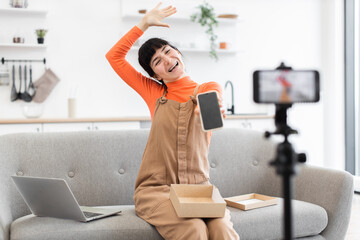 Caucasian young female blogger joyfully unboxes smartphone from cardboard box during online streaming. Expressive and cheerful, she captures moment using smartphone mounted on tripod.