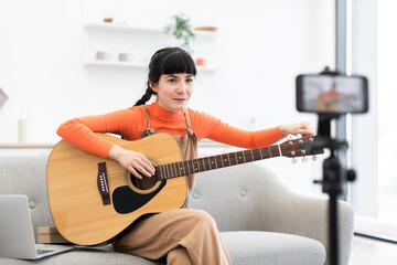 Caucasian female blogger recording online guitar lesson at home. She is sitting on couch with acoustic guitar, teaching through a camera. Bright setting suggests modern, creative environment.