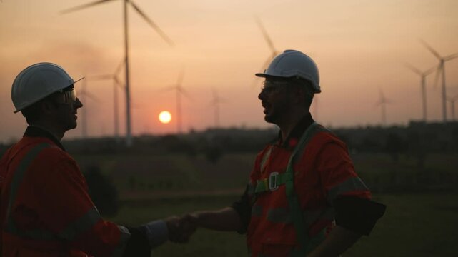 Technician workers discuss with handshake to work together at sunset outdoors at windmill farms. Renewable energy engineer working on wind turbine project. Engineer research environment green energy. 