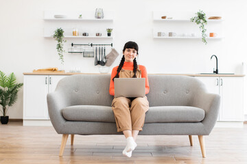Obraz premium Young Caucasian woman sitting comfortably on sofa with laptop in bright modern living room. Wearing orange sweater and brown overalls, she is focused on work-from-home environment.