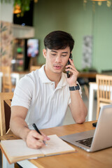 A focused Asian man is working remotely from a coffee shop, talking on the phone.