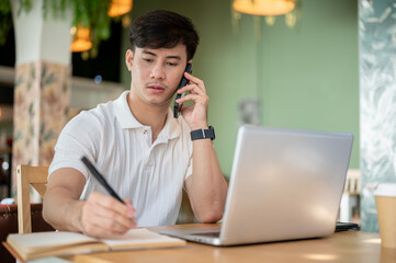 A focused Asian man is working remotely from a coffee shop, talking on the phone.