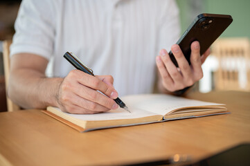 Fototapeta premium A cropped shot of an Asian man holding his smartphone while taking notes in his notebook.
