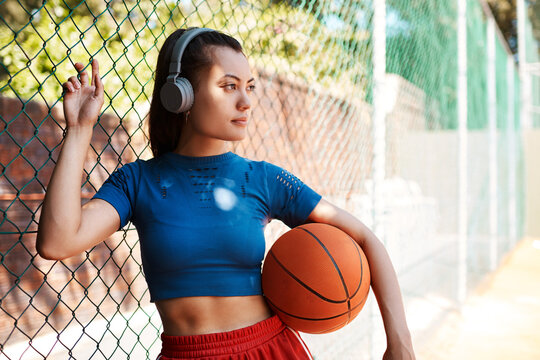 Woman, thinking and outdoor by fence as basketball player or athlete for challenge or exercise. Female person, planning and headphones in competition, match or game for idea, sport or training advice - Powered by Adobe