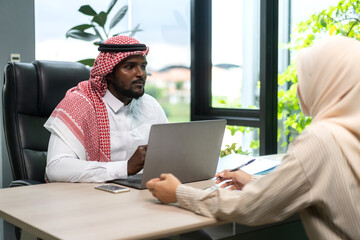 Professional arab middle eastern businessman in traditional attire working on laptop in modern office setting, discussing with female colleague, business communication and collaboration concept