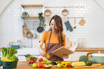 Asian young woman holds a book with online audience or simply enjoying the cooking process, health, fruit, freshness, and organic living, digital recipe platform, eating, healthy at kitchen