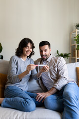 Happy couple looking at positive pregnancy test sitting on the sofa at home. Vertical image.