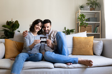 Smiling pregnant couple looking baby ultrasounds at home sitting on the couch.