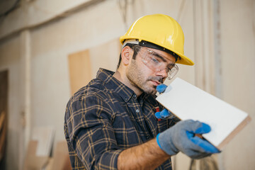 Construction worker in a yellow hardhat and safety glasses inspecting a piece of wood. Ideal for carpentry, woodworking, and quality control, highlighting craftsmanship in the workshop. labor day