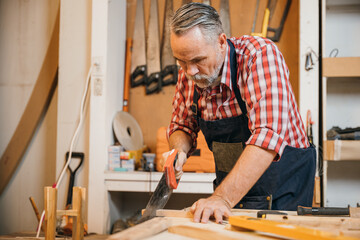 A carpenter in a red plaid shirt and blue overalls saws a piece of wood with a handsaw on a...