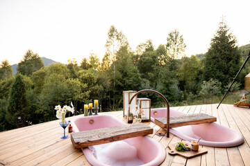 A tranquil setting with two pink outdoor bathtubs filled with bubbles, surrounded by nature. Candles and flowers add a touch of warmth, creating an ideal atmosphere for relaxation