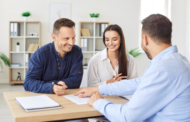 Obraz premium Cheerful family couple preparing to sign contract at realtor office. Professional real estate agent sitting at desk with happy clients, finalizing details of deal and showing where to put signature.