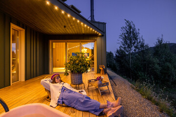 A woman relaxes on a deck under soft evening lights, wrapped in a cozy sweater and surrounded by...