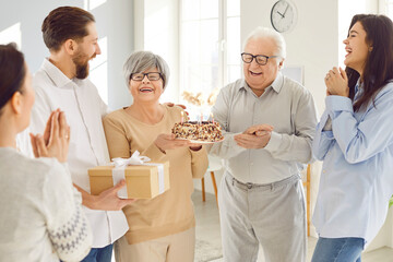 Happy senior woman celebrating birthday with her family standing in living room at home party. Smiling adult children with father congratulating old mother giving her cake and present gift box.