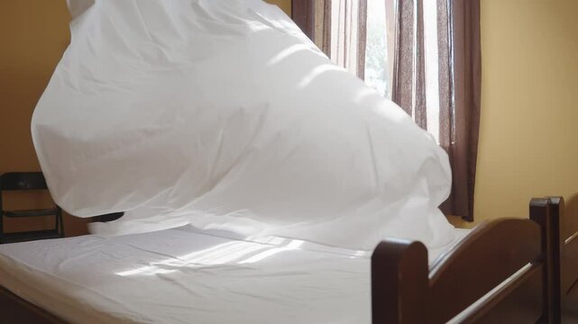 Housemaid covering the bed with a snow-white sheet. Female housekeeper changing bedding in a hotel room. Employee is cleaning a room in a hostel. Concept of quality service in the hotel business