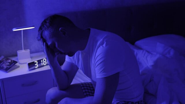 Man suffering from insomnia, sitting on bed near clock on bedside table. PTSD, post-traumatic stress disorder