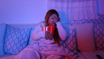 Scared woman covering herself in fear while watching horror movie sitting on sofa at night