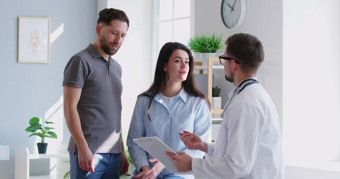 Portrait of a young happy couple talking with a male friendly family doctor therapist standing in medical clinic or hospital planning pregnancy. Health care and check up concept. 4k video.