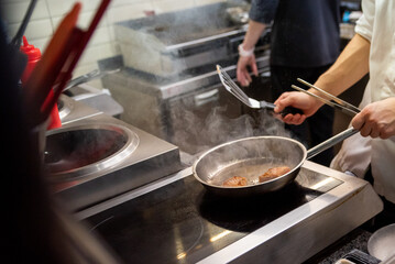 A chef in a professional kitchen sautés food in a pan on an induction stove, with steam rising. The scene captures the dynamic and focused atmosphere of culinary preparation
